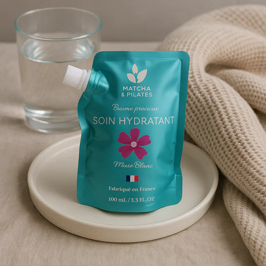 Soin hydratant Matcha & Pilates en sachet turquoise avec fleur rose, posé sur un plateau blanc à côté d'un verre d'eau et d'une couverture beige