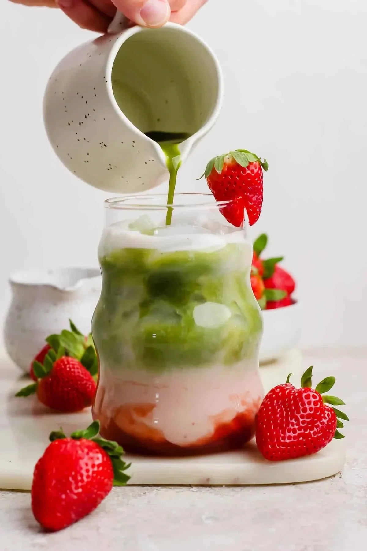 Verre de latte matcha fraise glacé avec fraises fraîches, versement de matcha vert dans un pot blanc